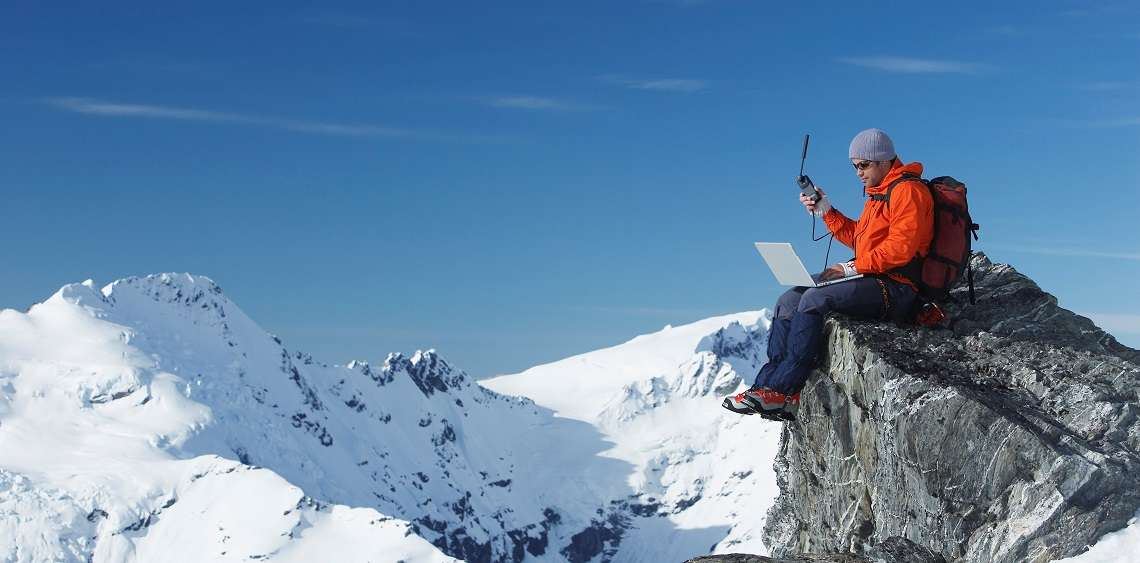 Person in roter Jacke sitzt auf Berggipfel mit Laptop und Antenne