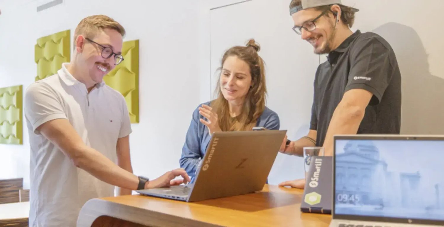 Drei SmartIT Mitarbeitende stehen an einem Tisch, lächeln und schauen auf einen Laptop